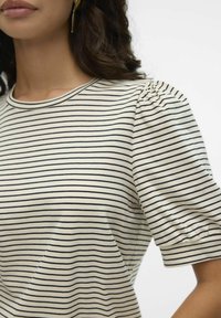 Γυναίκα που φοράει κοντομάνικη μπλούζα με μπεζ και μαύρες ρίγες, με φουσκωτά μανίκια και χρυσά ορθογώνια σκουλαρίκια.