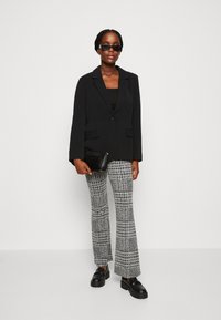 Blazer nero con un solo bottone e due tasche, abbinato a pantaloni grigi a quadri e mocassini neri con plateau. Clutch nera e occhiali da sole.