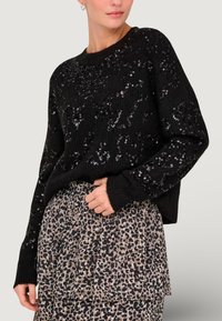 Pull en tricot noir orné de sequins brillants, avec des manches longues et un col rond, associé à une jupe beige à imprimé léopard.