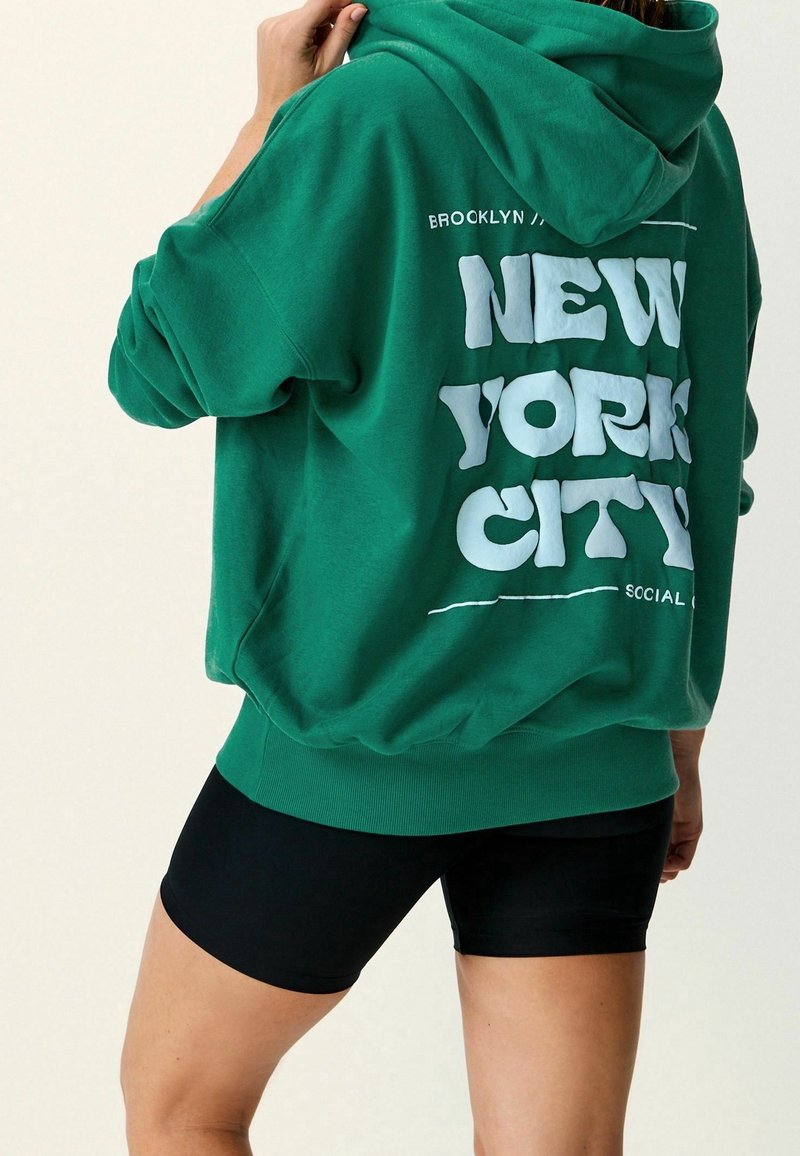 Persona con una sudadera verde que dice "New York City" en la parte de atrás y pantalones cortos de ciclismo negros, sosteniendo la capucha cerca de su cabeza.
