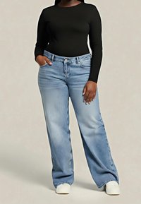 Kobieta ubrana w czarną bluzkę z długim rękawem, jasnoniebieskie szerokie dżinsy i białe buty, stojąca z jedną ręką w kieszeni, a drugą przy boku.