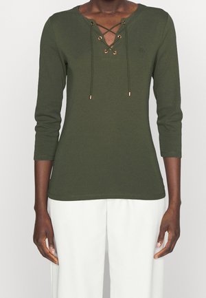 Person iført en olivengrøn langærmet top med snørelukning ved udskæringen og hvide bukser, stående mod en ensfarvet baggrund.