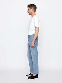 Bílá krátká tričko s kapsičkou na hrudi, spárované se světle modrými přímými džínami a černými nazouvacími botami, stojící v profilu.