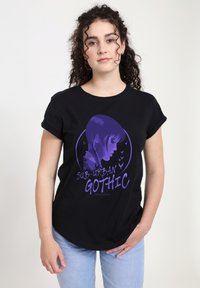 Mladá žena s kudrnatými vlasy, oblečená v černém tričku s fialovým profilem dívky s copánky, ptáky a nápisem "Sub-Urban Gothic," doplněná o světlé džíny.