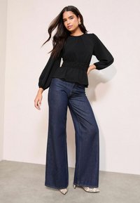 Maglia nera a maniche lunghe con peplum e vita arricciata abbinata a jeans a gamba larga blu scuro e tacchi con stampa pitonata su uno sfondo chiaro.