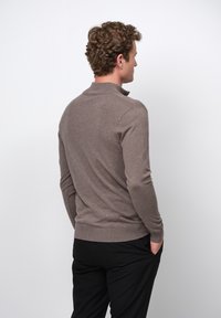 Brauner, taillierter Pullover mit hohem Kragen, strukturiertem Strickstoff und gerippten Bündchen, kombiniert mit schwarzen Hosen, von hinten betrachtet.