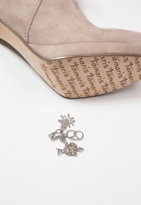 Beige suede shoe sole embossed with "Tamaris" repeated and silver charm with flower, pretzel, and heart with arrow on white background.