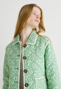Giacca trapuntata verde floreale con chiusura a bottoni, ampio colletto, piping a contrasto e tasche a toppa. Tessuto texturizzato con fiori bianchi.