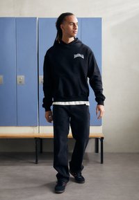 Black hooded sweatshirt with front pocket and logo, paired with dark jeans and black sneakers. Background features blue lockers.