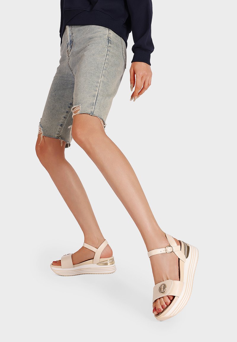 Sandali platform beige chiaro con una fascia superiore larga e cinturino alla caviglia regolabile. Presentano un accento metallico sulla suola e un design testurizzato.