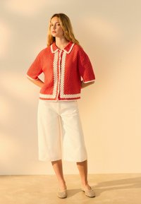 Cardigan corto a maglia all'uncinetto rosso con bordo bianco a onde, abbinato a pantaloni a palazzo bianchi e scarpe slip-on testurizzate.