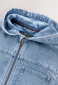 Veste en denim de couleur bleu clair, avec une fermeture éclair visible et une capuche. Comprend une poche frontale et une étiquette à l'intérieur. Tissu texturé.