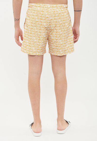 Pantaloni da bagno corti con un motivo ripetuto di logo giallo e bianco, vita elastica e tasche laterali. Indossati con scarpe slip-on bianche.