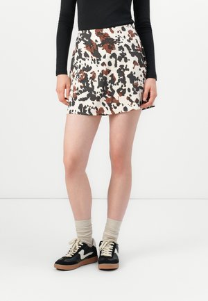 Kobieta ubrana w krótką spódniczkę z brązowo-czarnymi abstrakcyjnymi plamami, czarny top z długim rękawem, beżowe skarpetki i czarne trampki z białymi sznurowadłami.