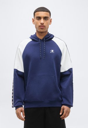 Mężczyzna ubrany w granatowo-białą bluzę Champion z kapturem i logo na piersi oraz wzorzyste paski na rękawach, stojący na jednolitym tle.