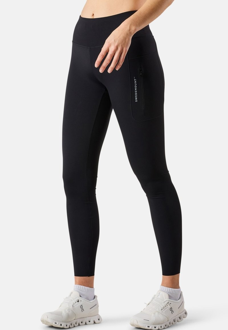 Leggings noirs au design ajusté, taille haute, poche zippée sur le côté et texture lisse. Associés à des chaussures de sport blanches.
