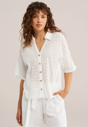 Frau mit lockigem Haar, die ein weißes Lochstickerei-Hemd mit Knöpfen, aufgekrempelten Ärmeln und passende Shorts trägt, steht mit einer Hand in der Tasche.