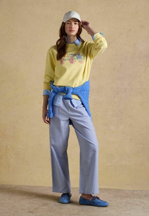 Mladá žena oblečená v žltej mikine s motívom auta, modrej košeli na gombíky, pruhovaných širokých nohaviciach, modrom svetri uviazanom v páse a modrých topánkach.