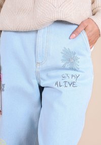Pantalon en denim bleu clair avec un design de fleurs brodées et le texte "RESTEZ EN VIE". Comprend des poches standard et des coutures contrastées.