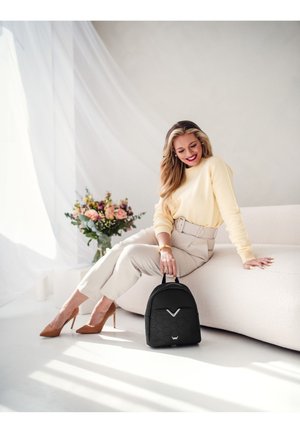 Černý batoh s hexagonální texturou, nastavitelnými popruhy a kovovým logem. Umístěn na jemném, světlém pozadí s květinovými akcenty.