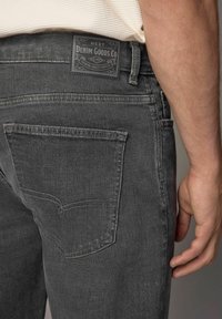 Sive traperice imaju stražnji džep sa šavovima i crnu kožnu oznaku na pojasu koja glasi "NEXT DENIM GOODS CO."