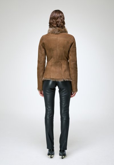 Personne aux cheveux ondulés portant une veste en daim marron avec bordure en fourrure et un pantalon en cuir noir, debout de dos sur un fond uni.