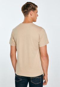 Beežist puuvillast T-särk lühikeste varrukatega ja ümar kaelus, ilma mustrita. Kandja on meesmodell, vaadatuna tagantpoolt.