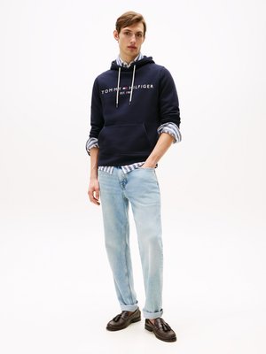 Hoodie marine avec logo blanc, jeans bleu clair et mocassins foncés. Superposé avec une chemise rayée, présentant un design décontracté et une texture douce.