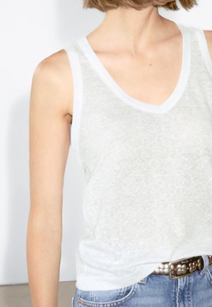 Persona che indossa una canottiera bianca a V scollata infilata in jeans blu con una cintura metallica borchiata, mostrata dal collo fino ai fianchi.