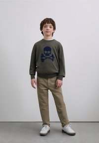 Sweater in olive green wool with a navy skull and crossbones design, paired with beige pants and white sneakers. Simple, casual fit.