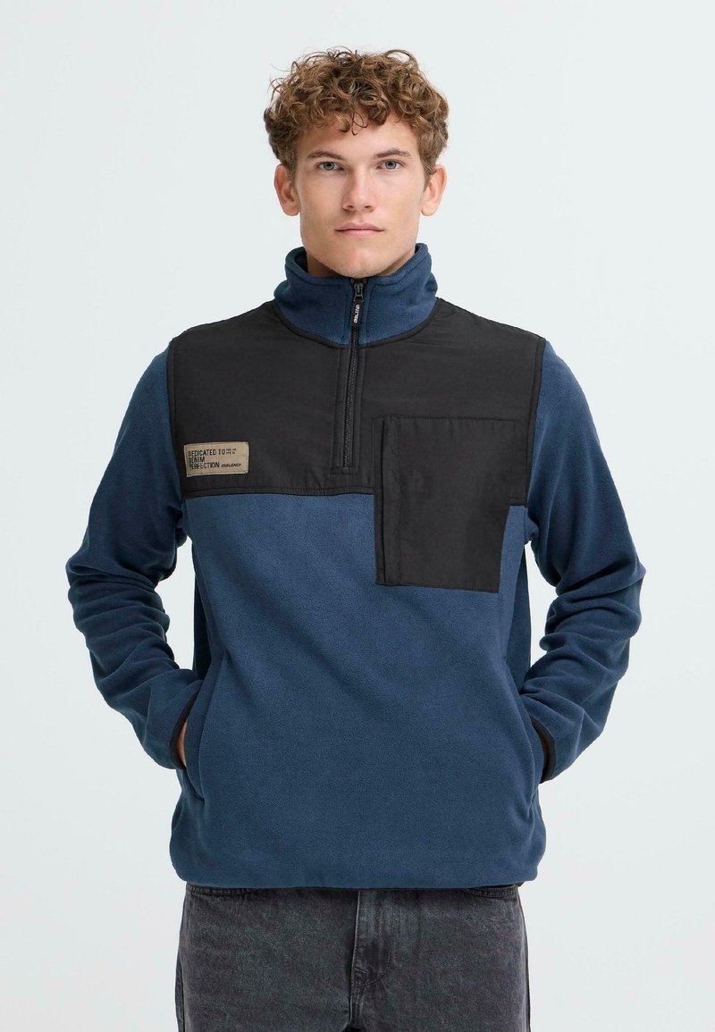 Pulóver de forro polar en azul marino con un panel negro en el pecho, cuello con cremallera media y bolsillo frontal que presenta una etiqueta. Suave al tacto y de corte relajado.