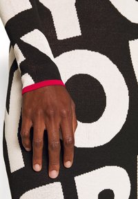 Robe en maille à motif noir et blanc avec un design de texte bien en évidence ; présente un accent de poignet côtelé rouge à la manche. Main posée sur le tissu.