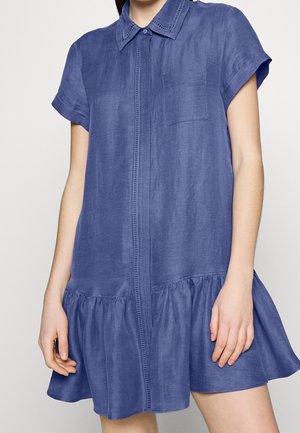 Vrouw draagt een blauw hemdjurk met korte mouwen, kraag, borstzak, geplooide zoom en verborgen knopen aan de voorkant tegen een effen achtergrond.