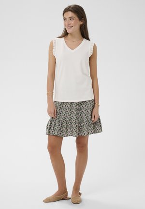 Mladá žena v bielom tielku bez rukávov s čipkovým lemom a kvetinovou sukňou stojaca bokom a usmievajúca sa.