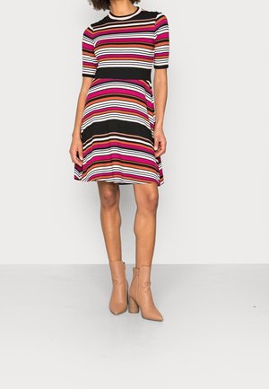 Femme portant une robe à manches courtes, longueur genoux, avec des rayures horizontales noires, blanches, roses, rouges et marron, associée à des bottines beige.