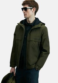 Veste imperméable vert olive avec capuche, deux poches avant et fermeture éclair. Portée sur une chemise noire, tenue d'une main.