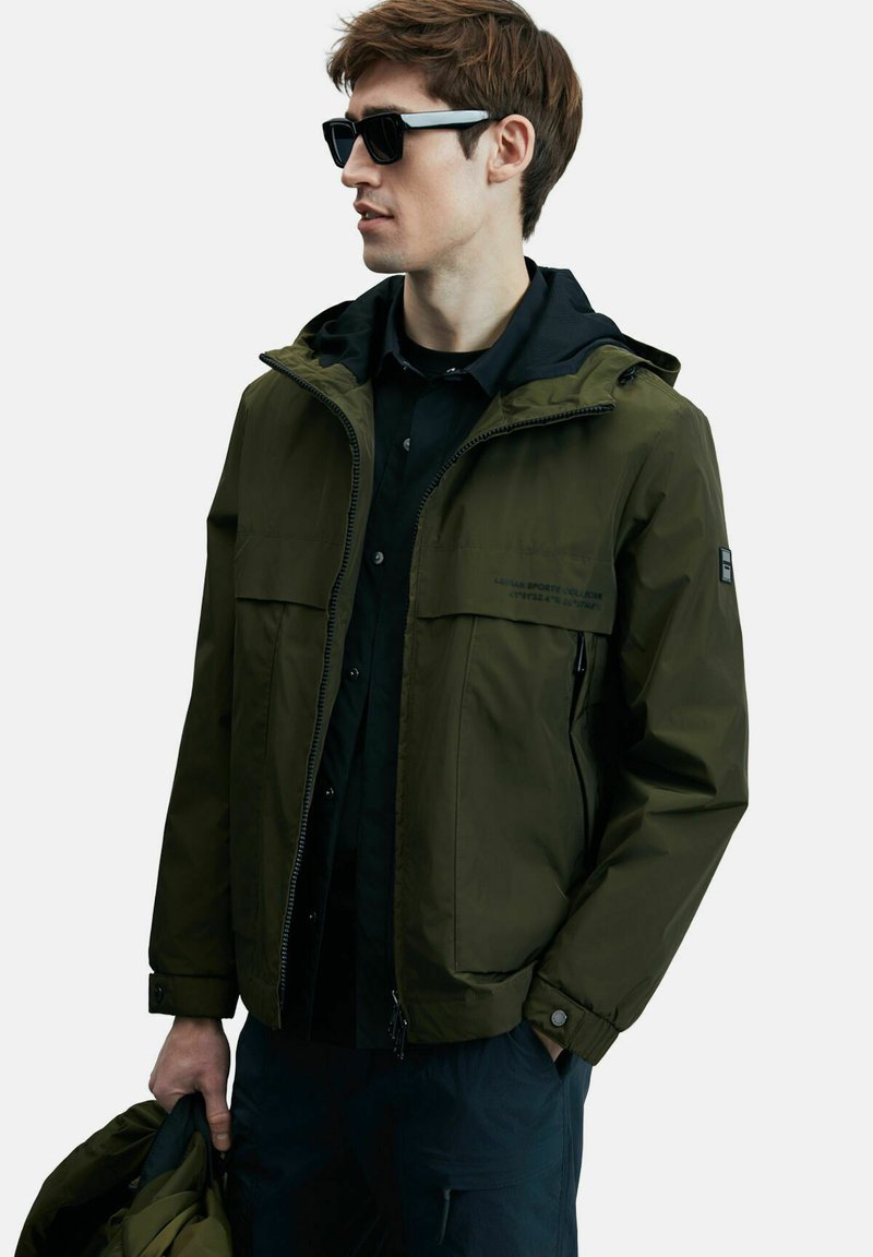Veste imperméable vert olive avec capuche, deux poches avant et fermeture éclair. Portée sur une chemise noire, tenue d'une main.