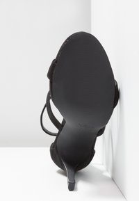 Sandale noire à talon haut reposant sur son talon, avec des brides visibles et une semelle lisse sur un fond blanc en coin.