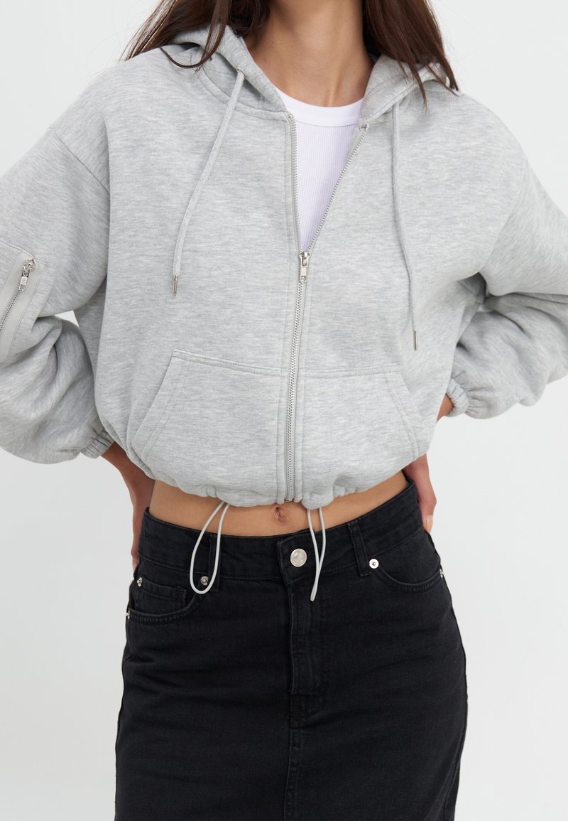 Kobieta ubrana w jasnoszary krótką bluzę z zamkiem błyskawicznym na białej koszulce, w połączeniu z czarną dżinsową spódnicą o wysokim stanem. Ręce na biodrach.
