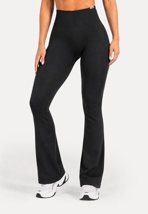 Kvinde iført sorte flared leggings med høj talje og hvide sportssko, stående med benene let fra hinanden på hvid baggrund.