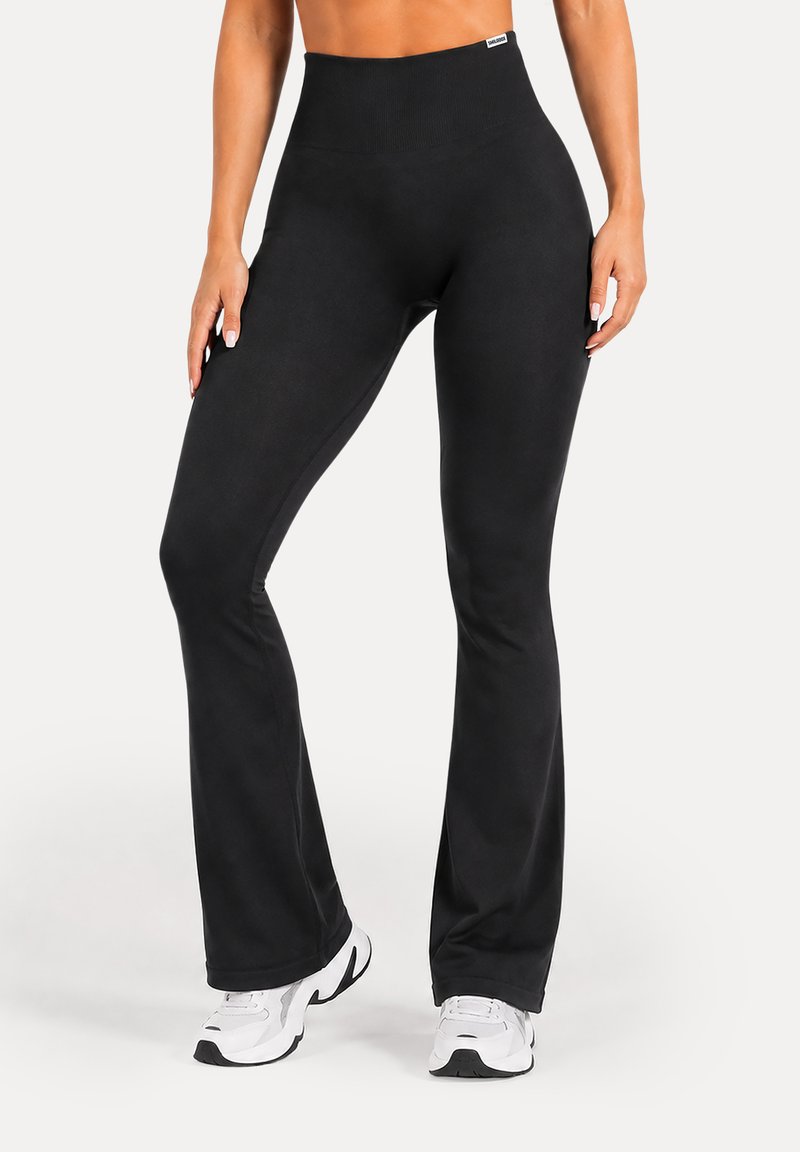 Femme portant un legging noir évasé taille haute et des chaussures de sport blanches, debout, jambes légèrement écartées sur fond blanc.