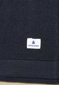 Mörk marinblå strukturerad tyg med ribbat manschett, med en vit etikett som visar varumärkesnamnet "JACK&JONES" i blå bokstäver.