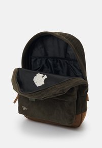 Sac à dos en velours côtelé vert foncé avec un rabat en toile, logo brodé, base en suède marron et un intérieur spacieux avec doublure noire.