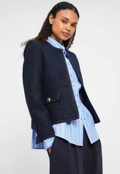 Giacca corta in tweed blu navy con collo rotondo, dettagli in bottone dorato sulle tasche, abbinata a una camicia a righe blu chiaro sotto.