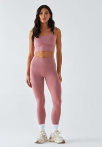 Różowy sportowy biustonosz i dopasowane legginsy wykonane z gładkiego, rozciągliwego materiału. Całość dopełniają białe skarpetki do kostek i lekkie sneakersy.