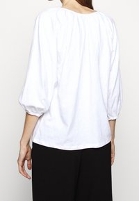 Biała bluzka z trzema ćwierć rękawami puff, teksturowana tkanina, plisowany dekolt i luźny krój, w komplecie z czarnymi spodniami.