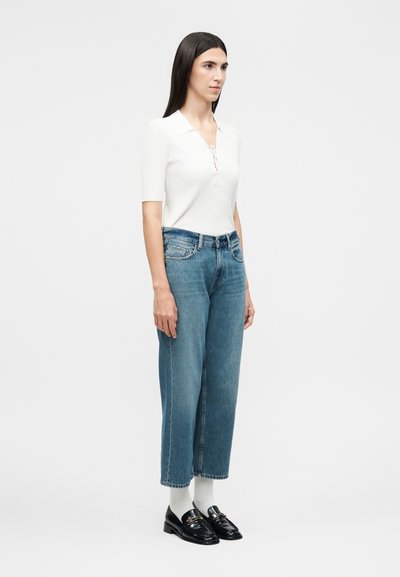 Γυναίκα στέκεται, φοράει λευκό πλεκτό τοπ με κοντά μανίκια, μπλε φαρδιά cropped τζιν, λευκές κάλτσες και μαύρα δερμάτινα loafers σε απλό φόντο.