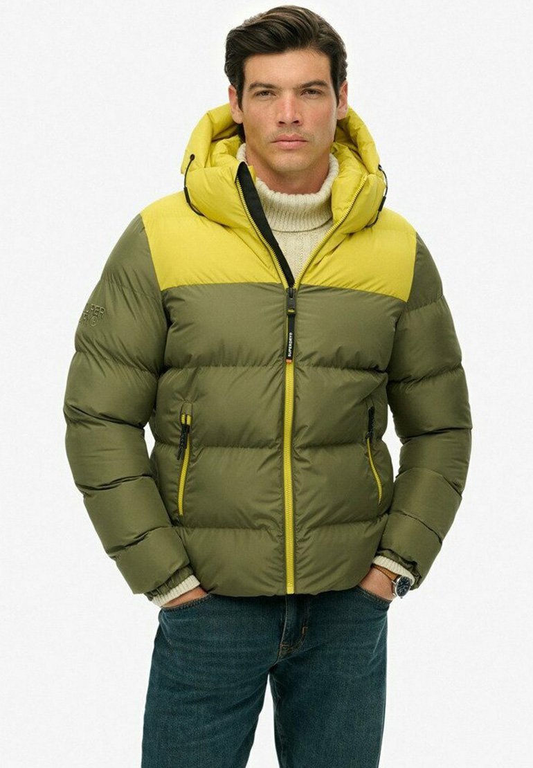Chaqueta acolchada verde y amarilla con capucha, que cuenta con bolsillos con cremallera y puños acanalados, llevada sobre un suéter crema y jeans azules.