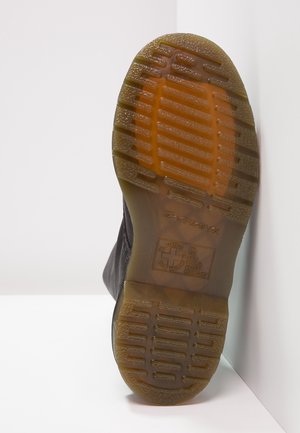 Semelle de chaussure avec bande de roulement en caoutchouc brun translucide présentant des rainures rectangulaires et un texte en relief, inclinée sur fond blanc aux angles.