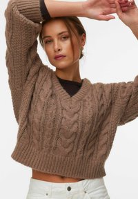 Jeune femme aux cheveux châtain clair portant un pull en maille épaisse marron et un pantalon blanc, levant les bras au-dessus de la tête sur un fond blanc.